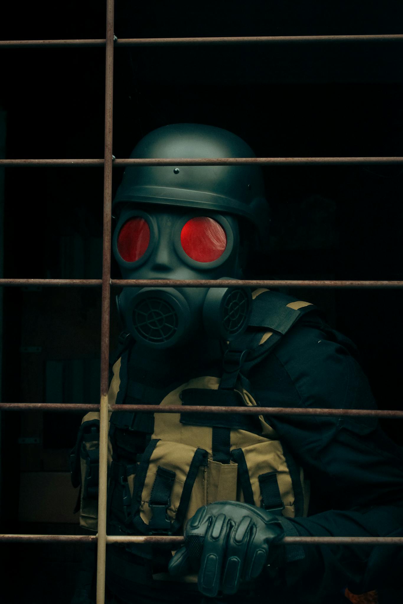 A person wearing a gas mask and tactical gear, standing behind metal bars.